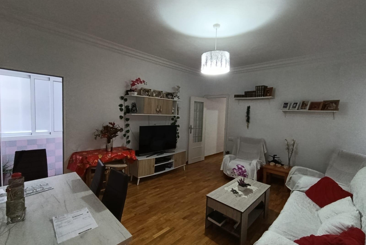 Cozy living room with white sofa and red cushions, wooden floor, and a white coffee table; TV on a white entertainment unit with shelves and plants nearby. A dining table on the left and a bright ceiling light illuminate the room.