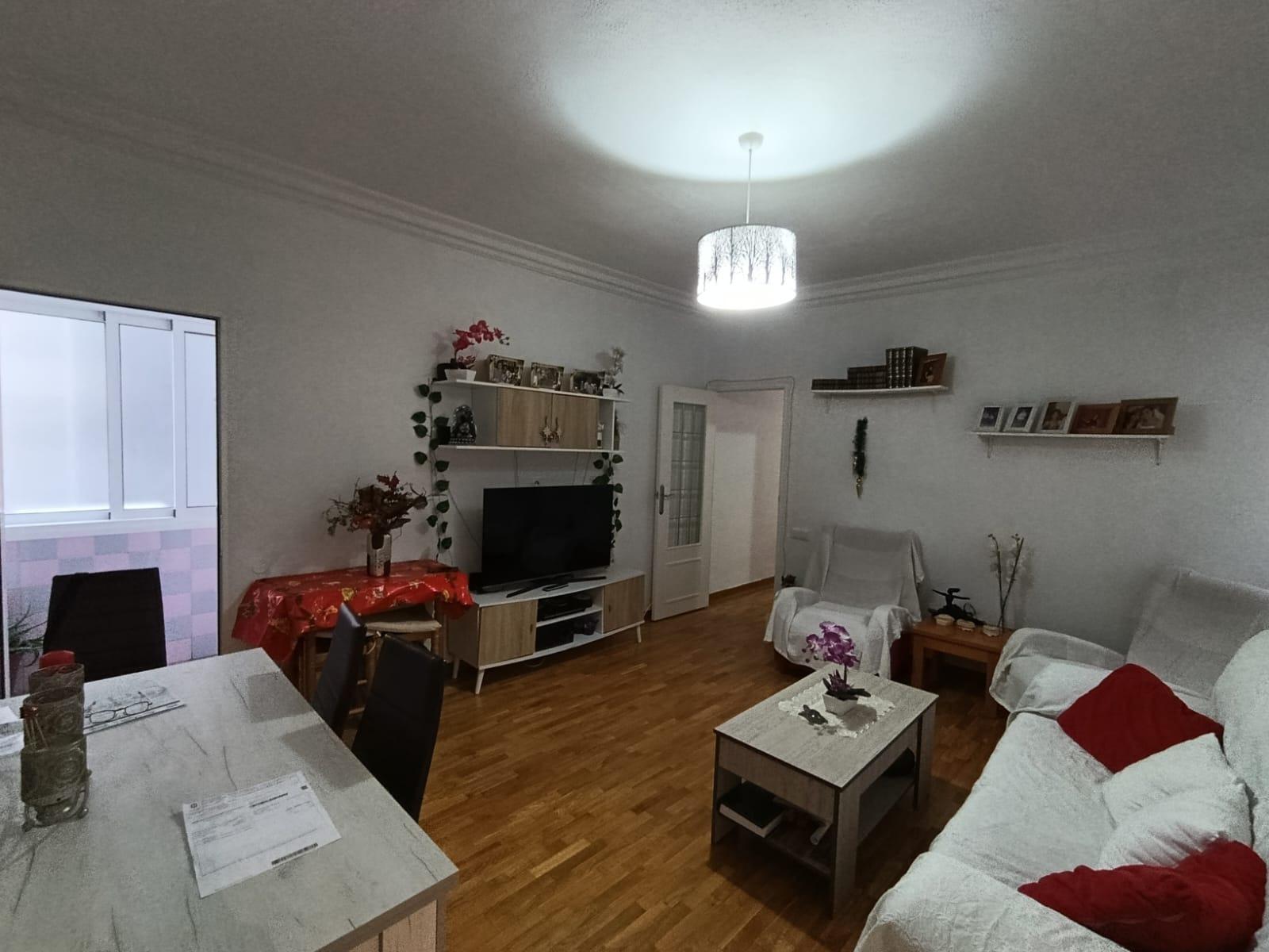 Cozy living room with white sofa and red cushions, wooden floor, and a white coffee table; TV on a white entertainment unit with shelves and plants nearby. A dining table on the left and a bright ceiling light illuminate the room.