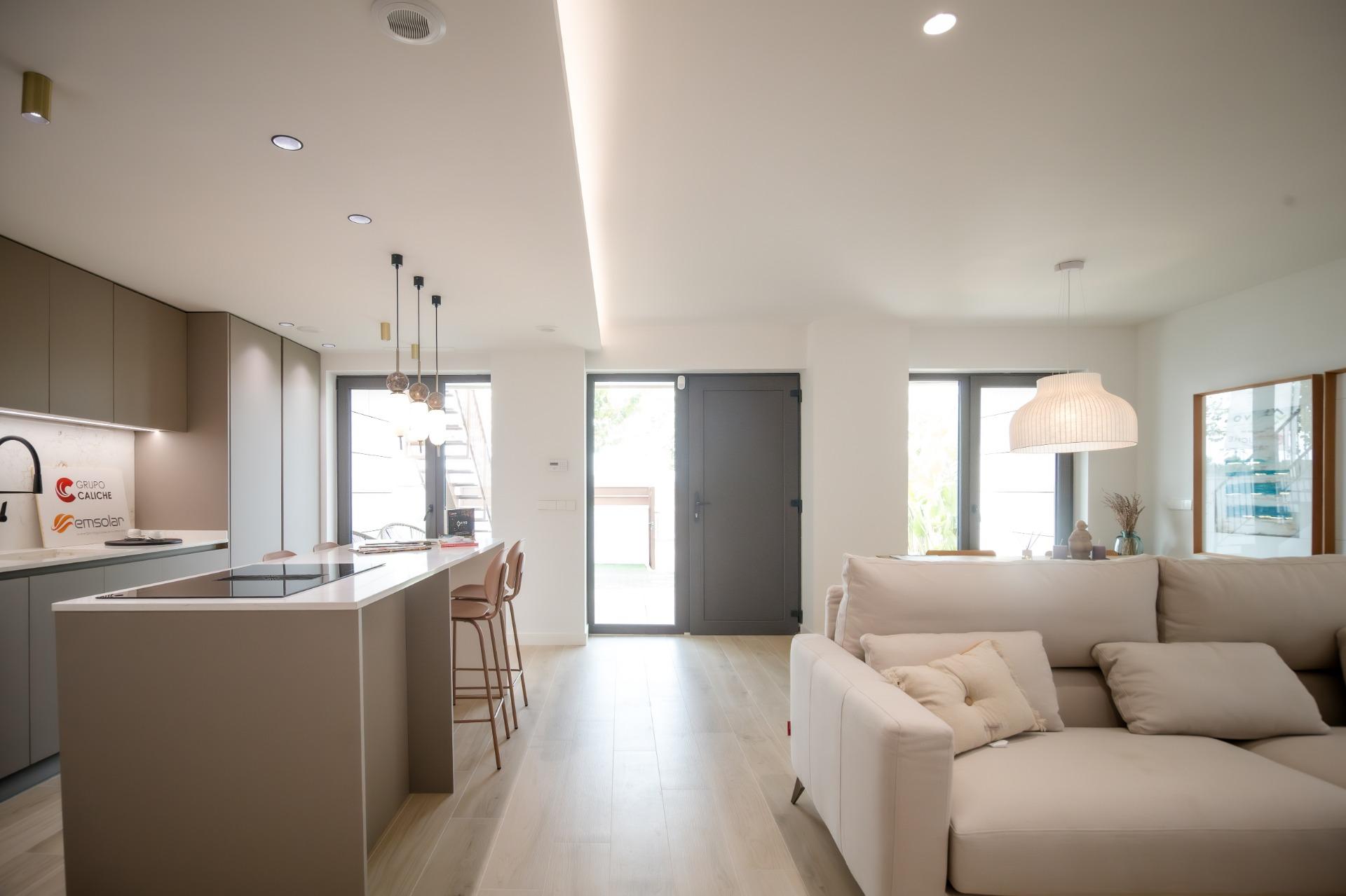 Open-plan kitchen and living area with taupe cabinets, white island, pendant lights, and a beige sectional sofa under bright natural light.