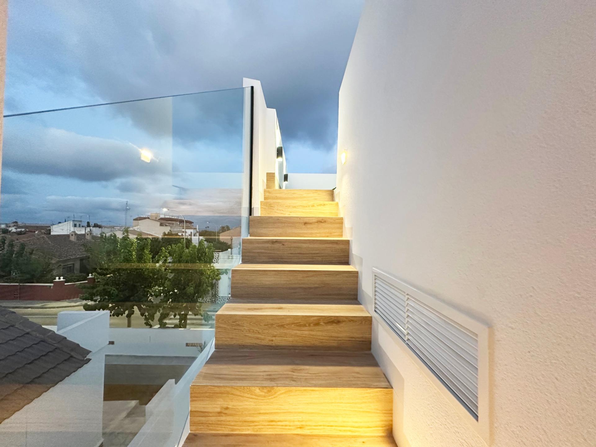 Wooden open-riser staircase with a glass railing, white walls, ascending to a bright upper landing; outside shows a cloudy blue sky and houses.