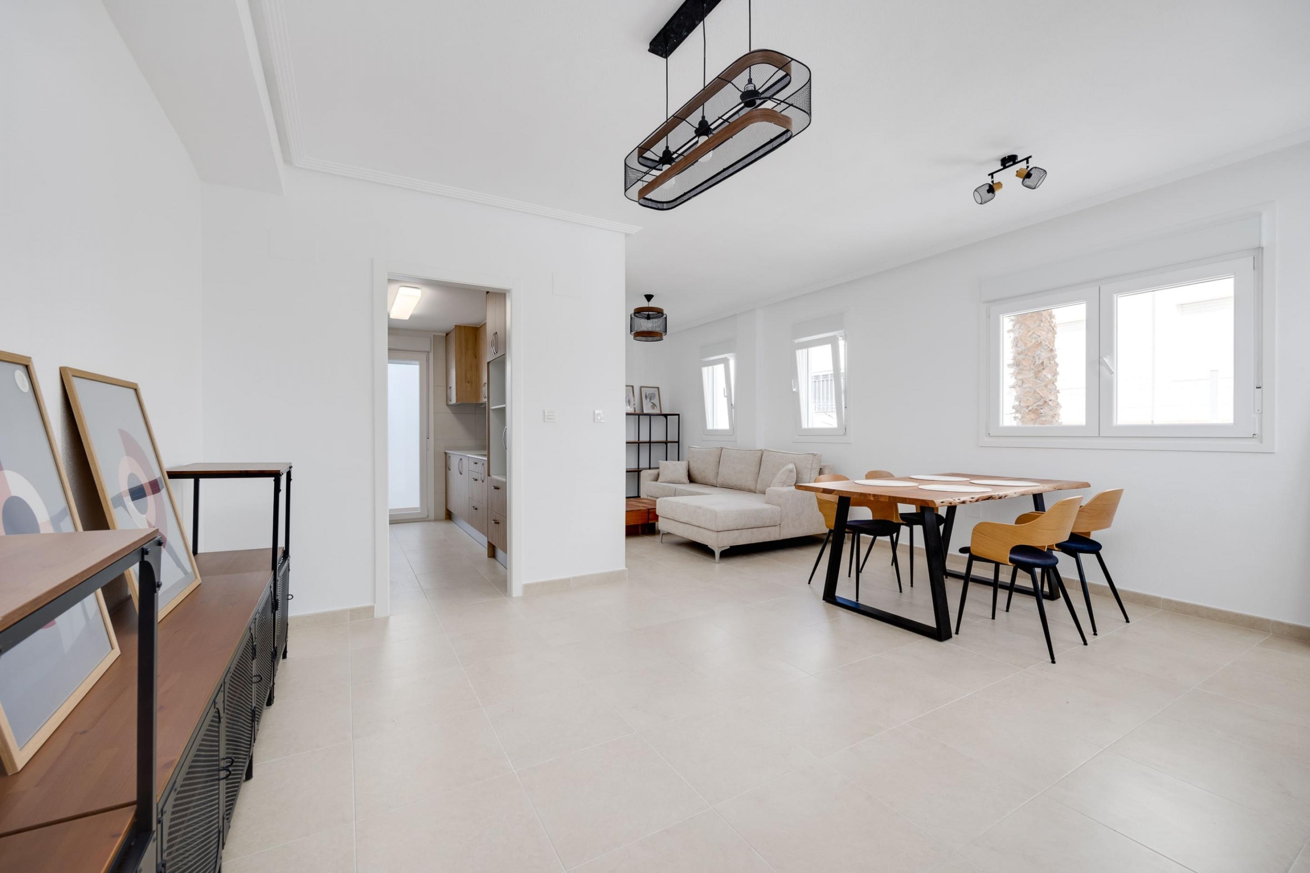 Bright open-plan living and dining area with a beige sofa, wooden dining table and chairs, and large white-framed windows.