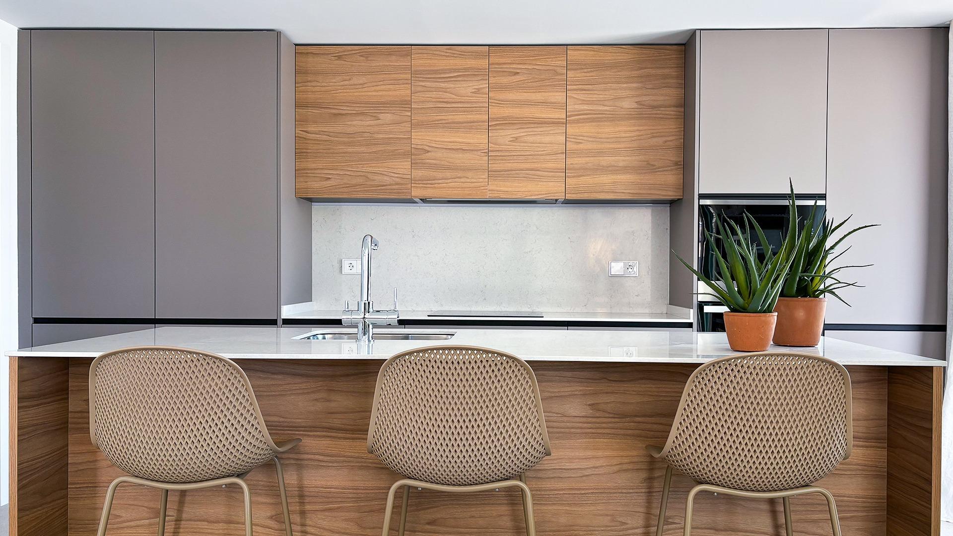 Modern kitchen with wood upper cabinets, grey tall cabinetry, marble backsplash, and a white island with three woven chairs.