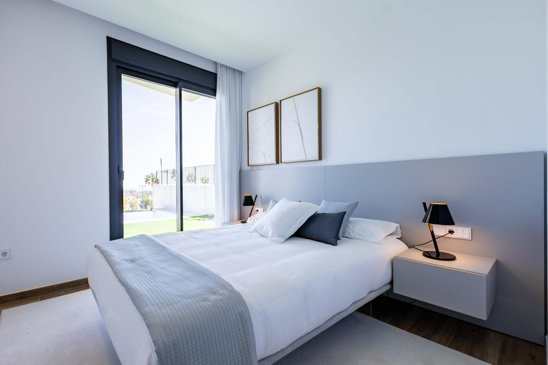 Modern bedroom with a low gray headboard, white bedding, and two black bedside lamps; a large window with sheer curtain opens to a balcony outside.