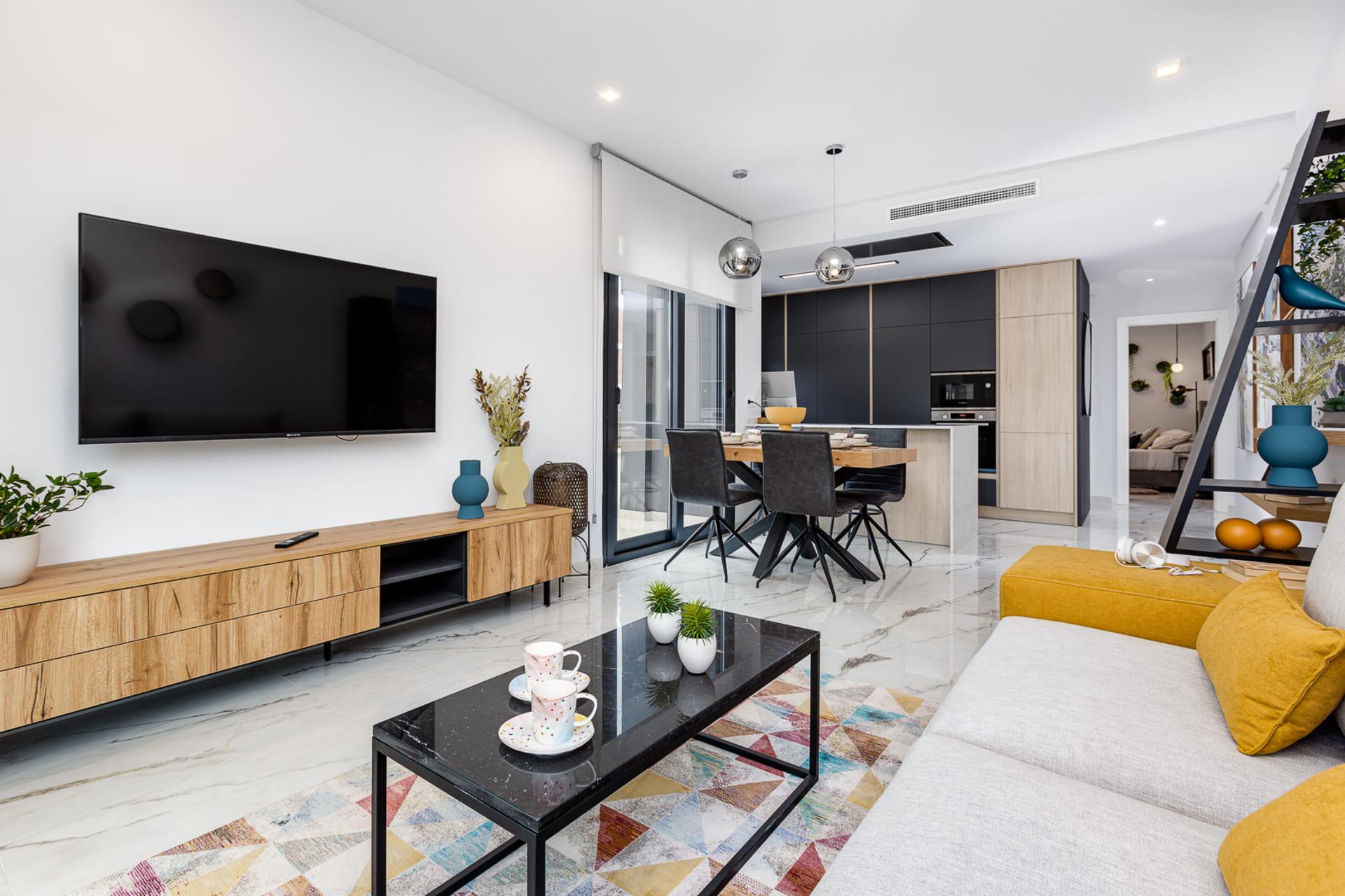 Open-plan living area with a wall-mounted flat-screen TV, wooden media console, and a gray-and-yellow sofa. The kitchen is in the background.