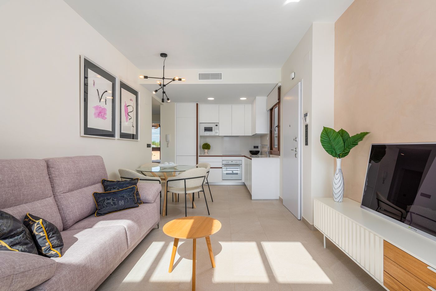 Open-plan living area with a pale pink sofa, decorative cushions, round glass dining table, and a white modern kitchen in the background under a black starburst light fixture.