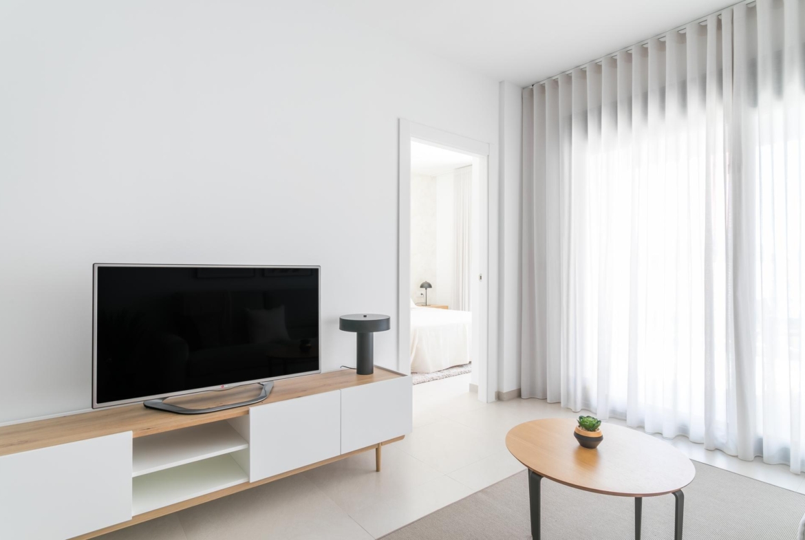 Minimalist living room with a flat-screen TV on a white-and-wood media console and a round coffee table near sheer white curtains.