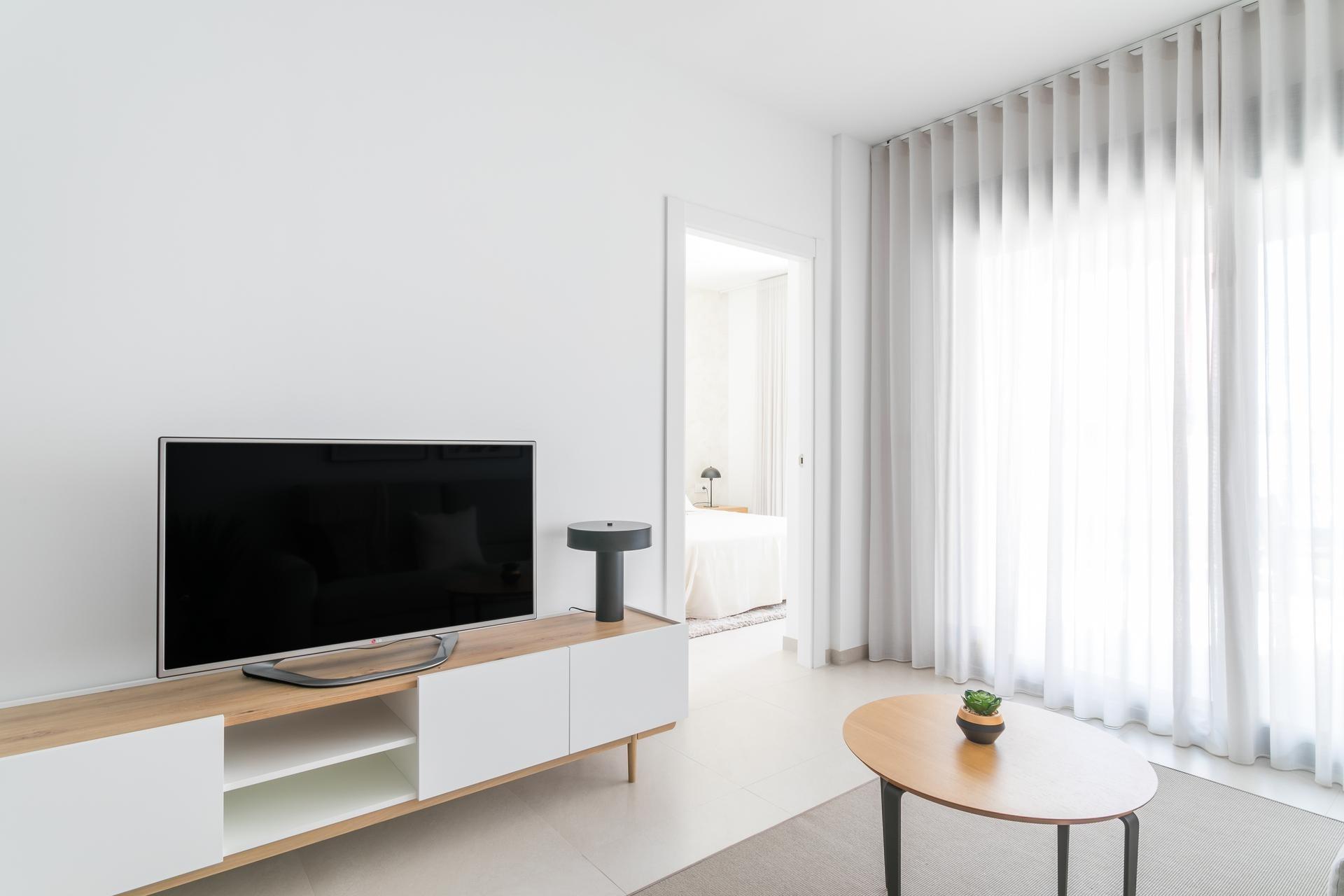 Minimalist living room with a flat-screen TV on a white-and-wood media console and a round coffee table near sheer white curtains.