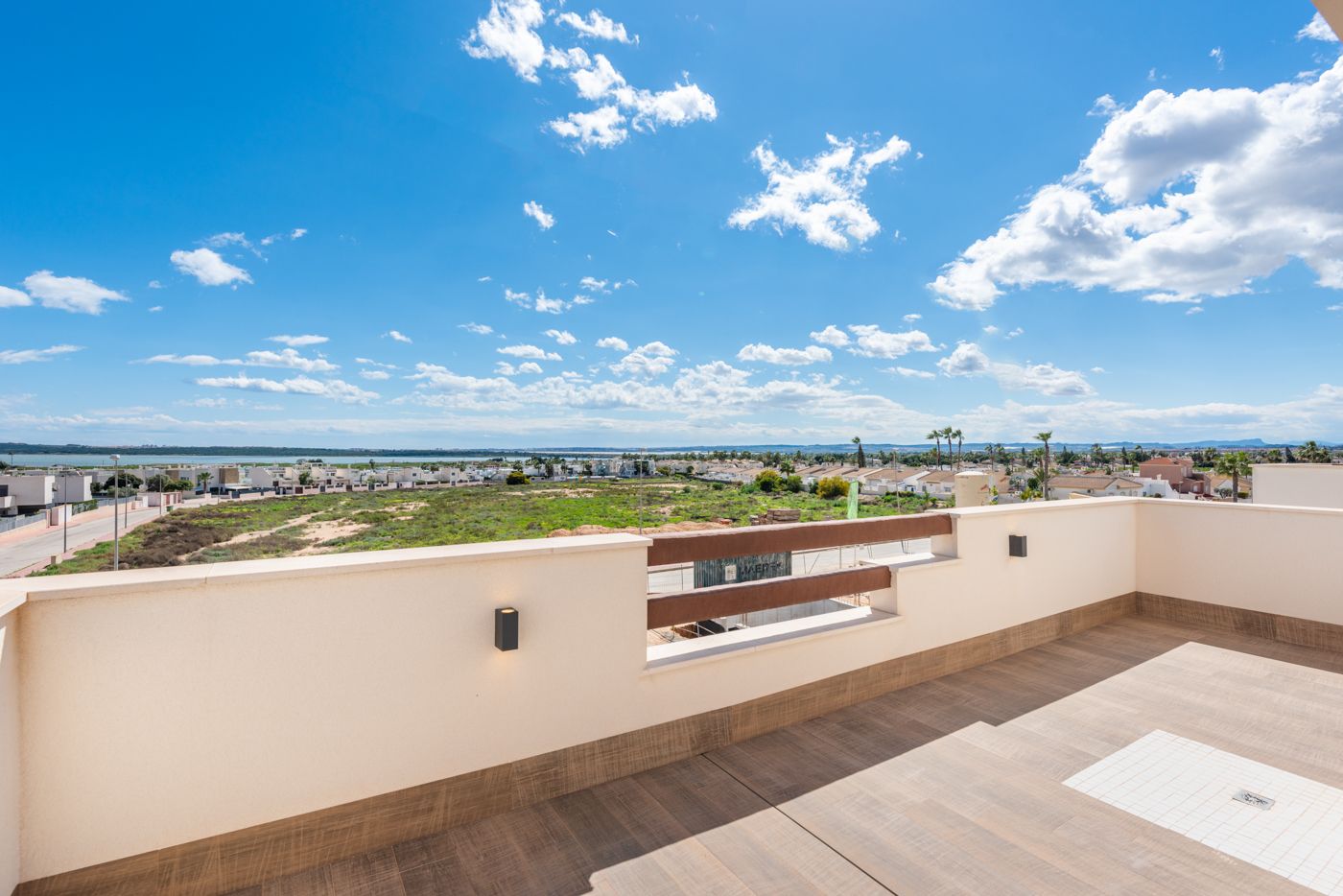 Rooftop terrace with beige walls and wooden railing, overlooking a desert landscape and distant houses under a clear blue sky with scattered clouds.