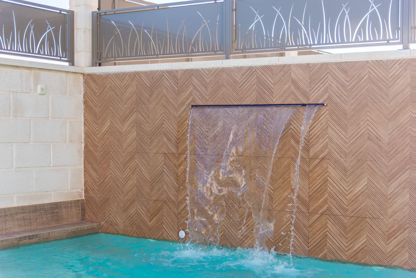 Water feature: a sheet of water cascading from a narrow spout into a turquoise pool, set in a brown herringbone tiled wall.