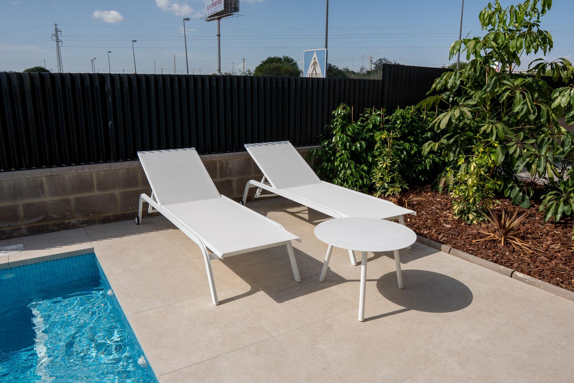 Two white pool lounge chairs and a round side table on a tan-tiled patio, with a tall black privacy fence and green shrubs nearby under a blue sky, near a pool edge.