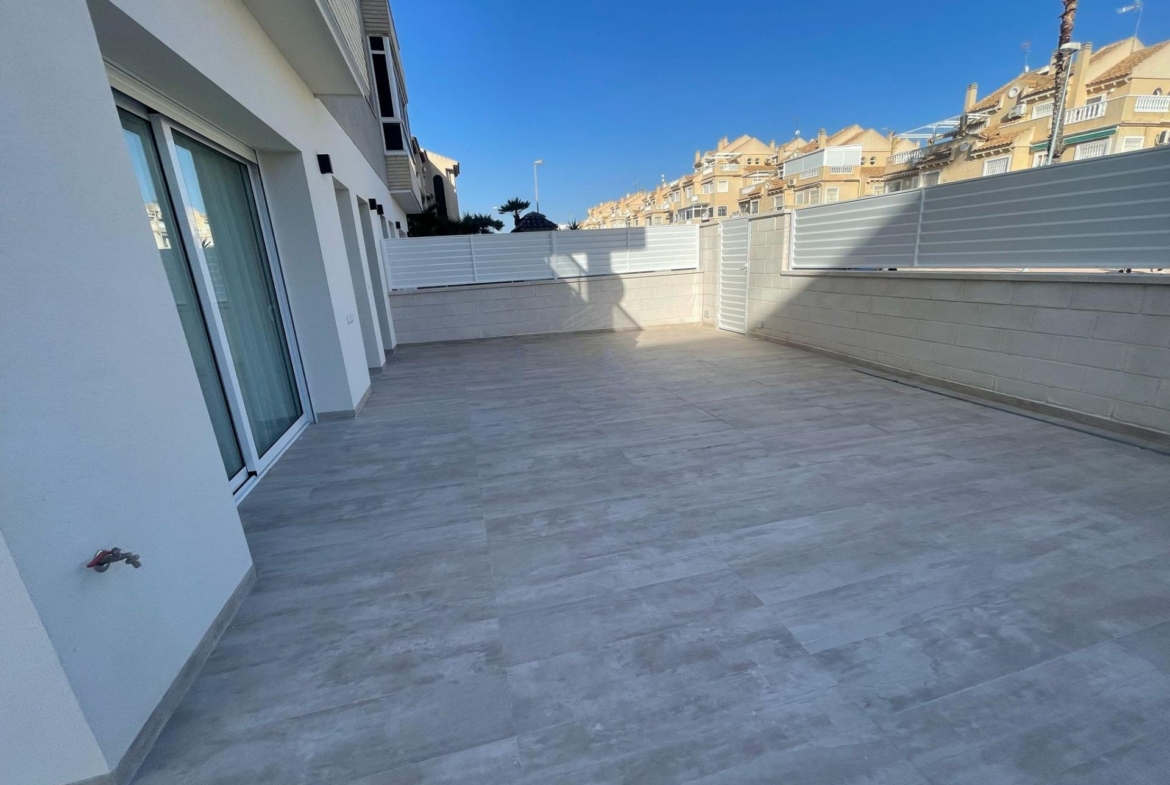 Outdoor terrace with gray tiled floor, sliding glass doors, white walls, and a white privacy fence; urban buildings visible against a clear blue sky