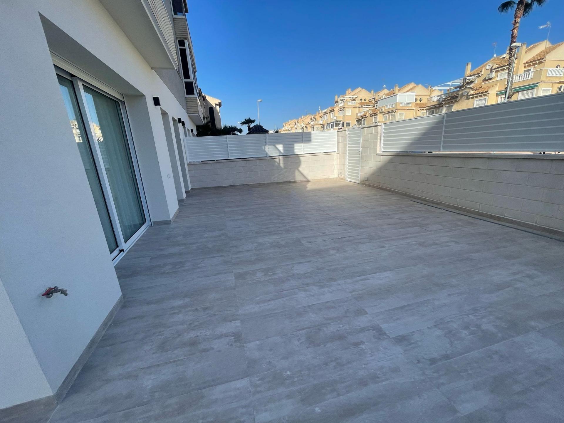 Outdoor terrace with gray tiled floor, sliding glass doors, white walls, and a white privacy fence; urban buildings visible against a clear blue sky