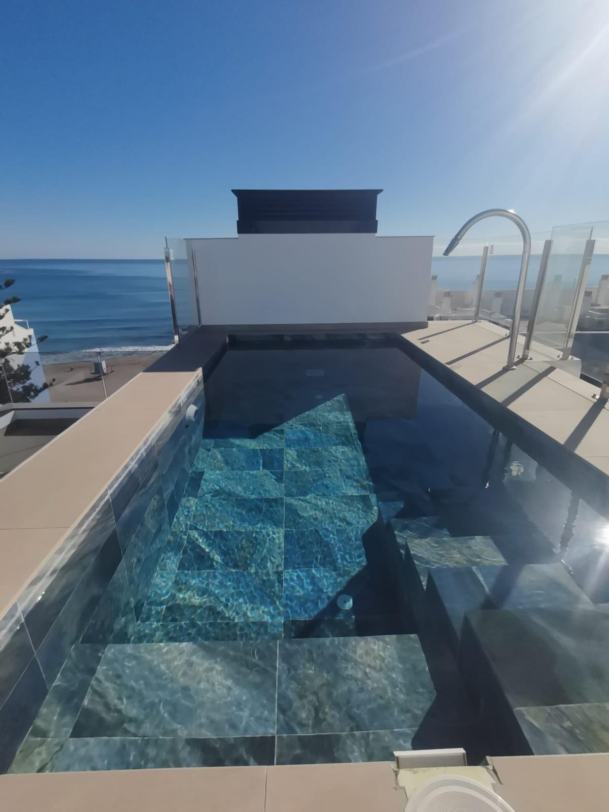 Rooftop infinity pool with blue tiled interior, glass railing, and ocean view under a bright blue sky.
