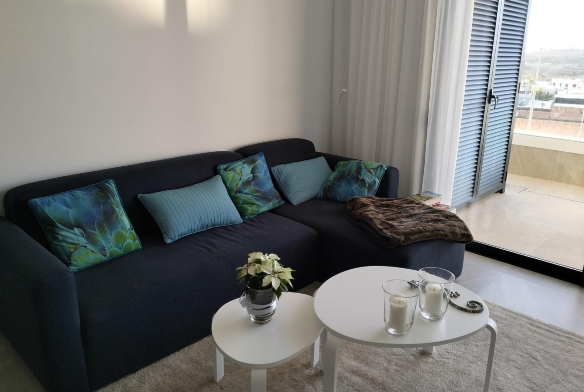Living room with a dark blue sectional sofa, teal and blue cushions, two white nested coffee tables, a small plant, and candles; glass doors with blinds opened to a balcony view.