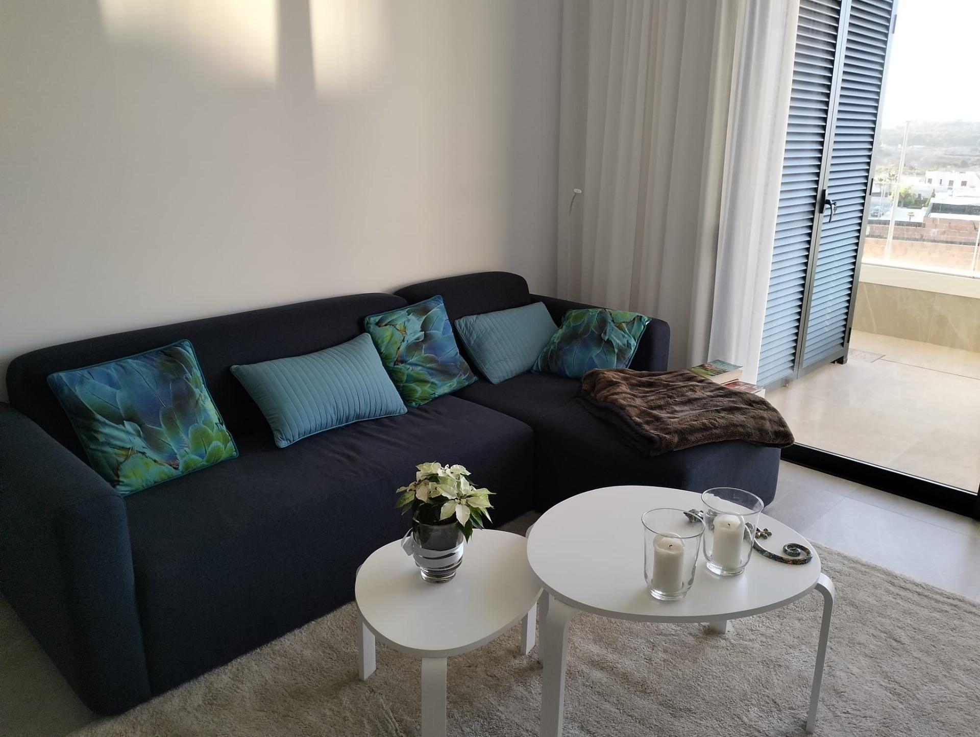 Living room with a dark blue sectional sofa, teal and blue cushions, two white nested coffee tables, a small plant, and candles; glass doors with blinds opened to a balcony view.