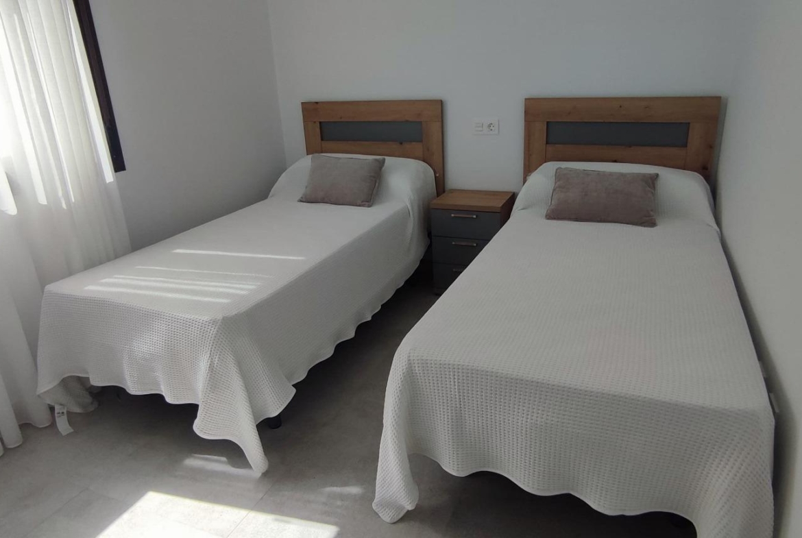 Two single beds with white textured bedspreads and gray pillows, separated by a small gray nightstand in a bright, minimalist bedroom.