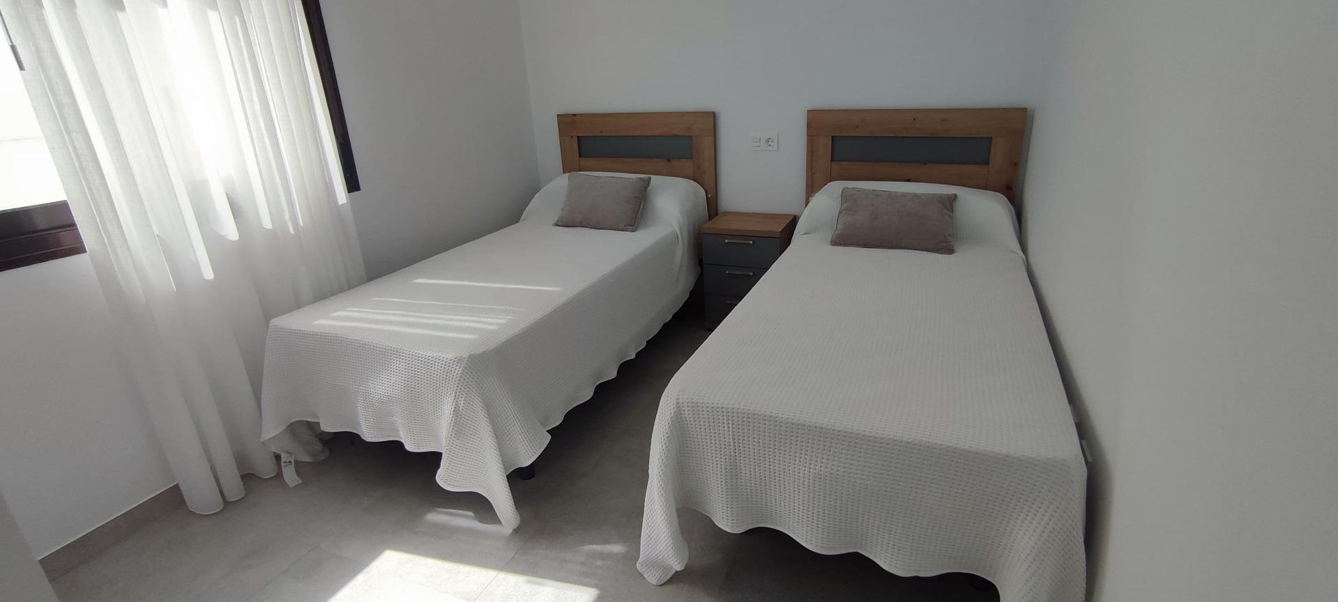 Two single beds with white textured bedspreads and gray pillows, separated by a small gray nightstand in a bright, minimalist bedroom.