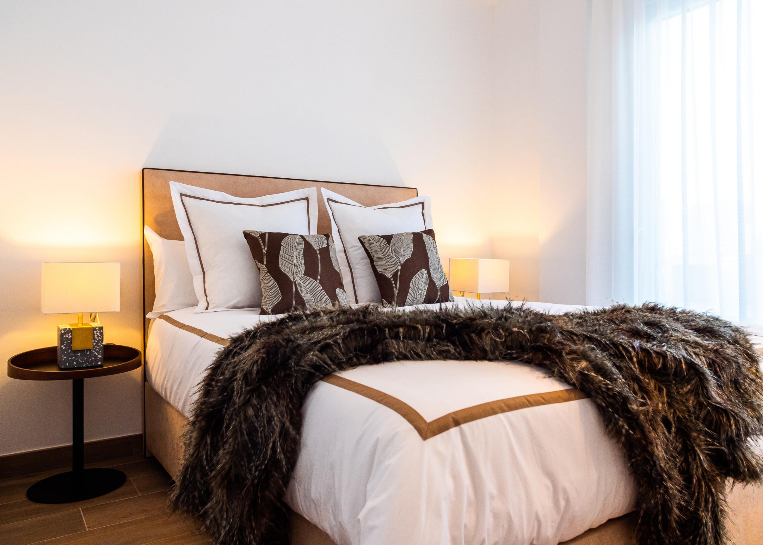 Bed with white linens and brown-accent pillows, a fluffy fur throw at the foot, and warm bedside lamps on round tables in a bright bedroom.