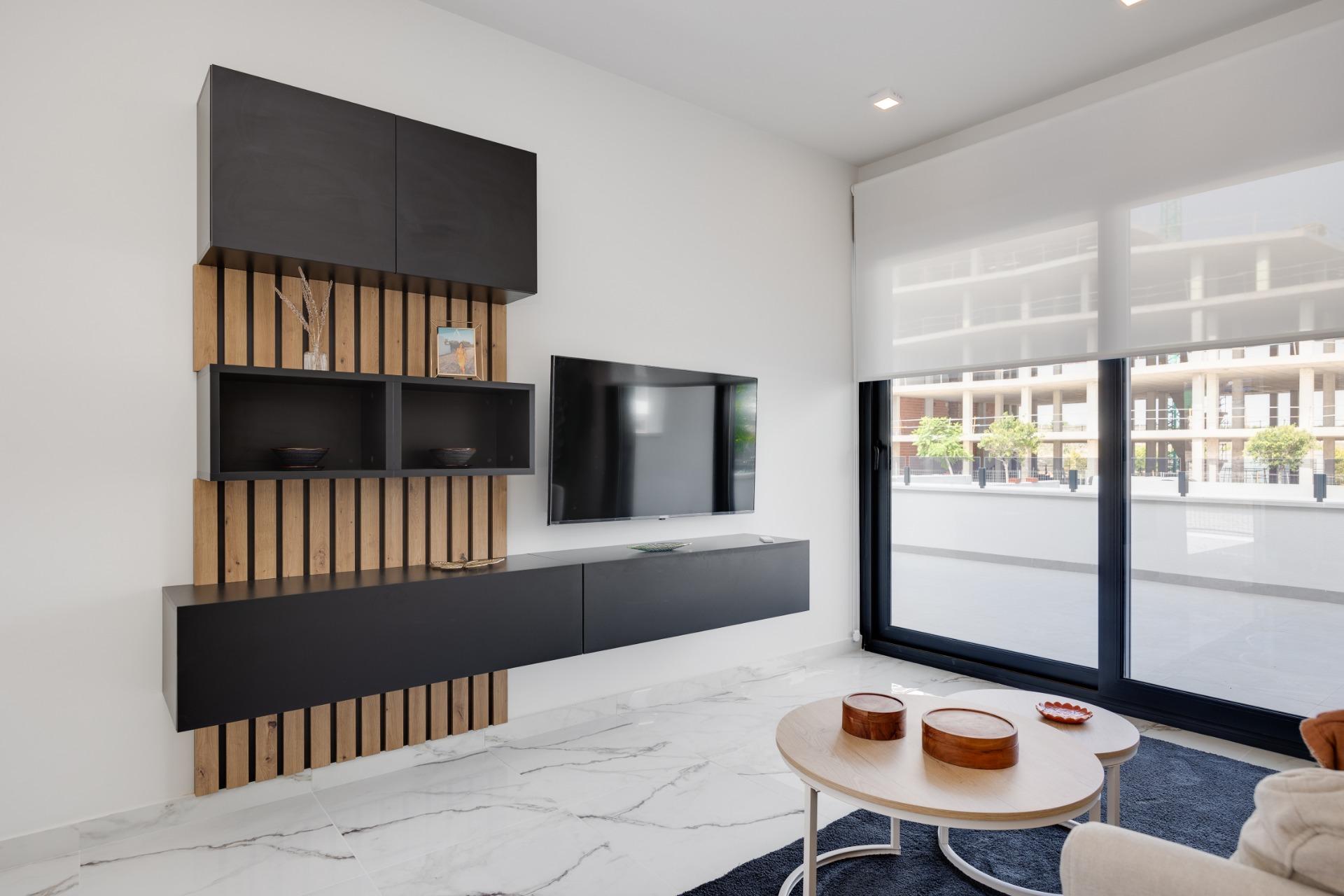 Modern living room with a wall-mounted black storage unit, wooden slats, and a flat-screen TV by large glass windows with white roller blinds.
