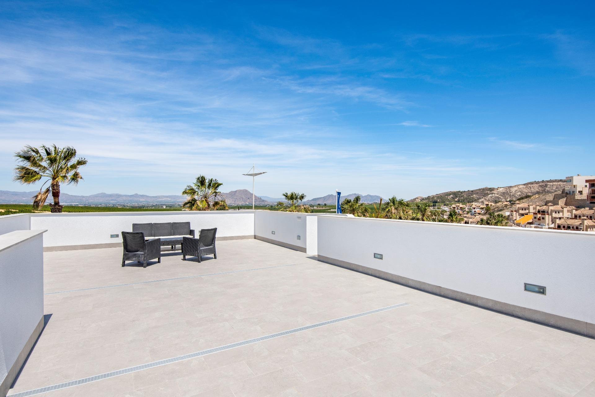 Rooftop terrace with a wicker seating set and white walls, palm trees, and distant hills under a clear blue sky.