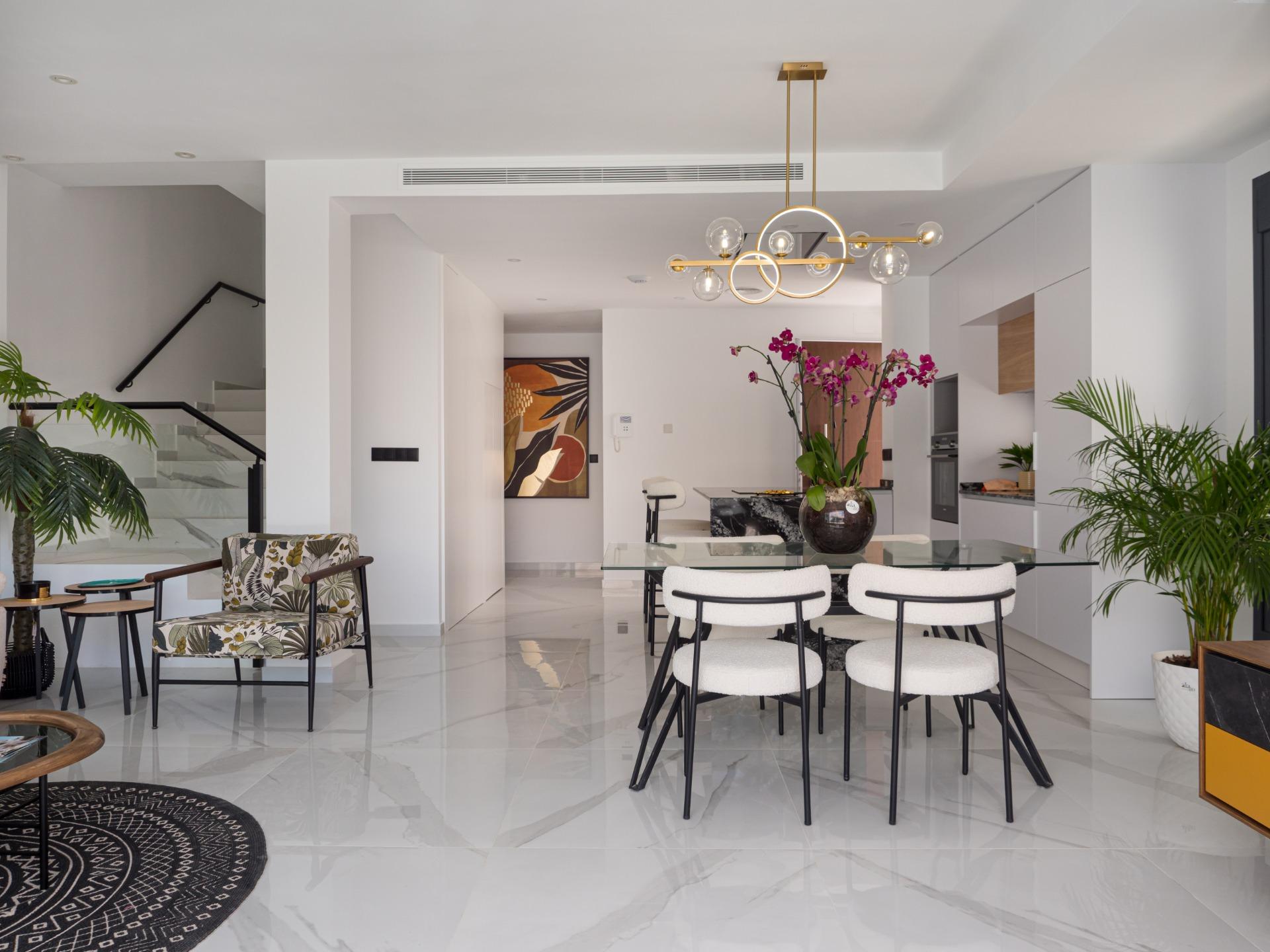 Open-plan dining area with a glass table, six white upholstered chairs, a gold chandelier, and large indoor plants; kitchen in the background.