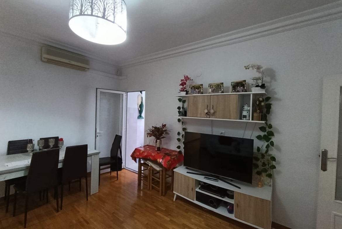 Living room interior with a TV on a white-wood stand, shelves, and climbing plants along the cabinet. A red tablecloth table sits near the door to the left.