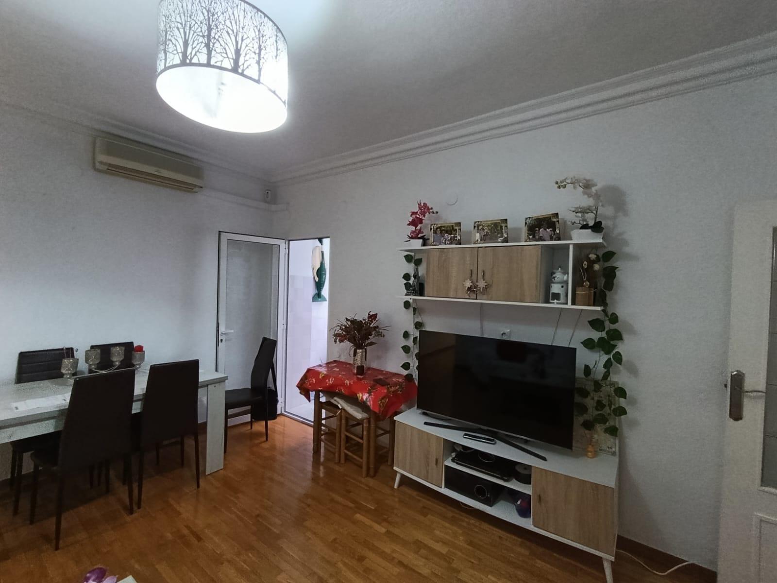 Living room interior with a TV on a white-wood stand, shelves, and climbing plants along the cabinet. A red tablecloth table sits near the door to the left.