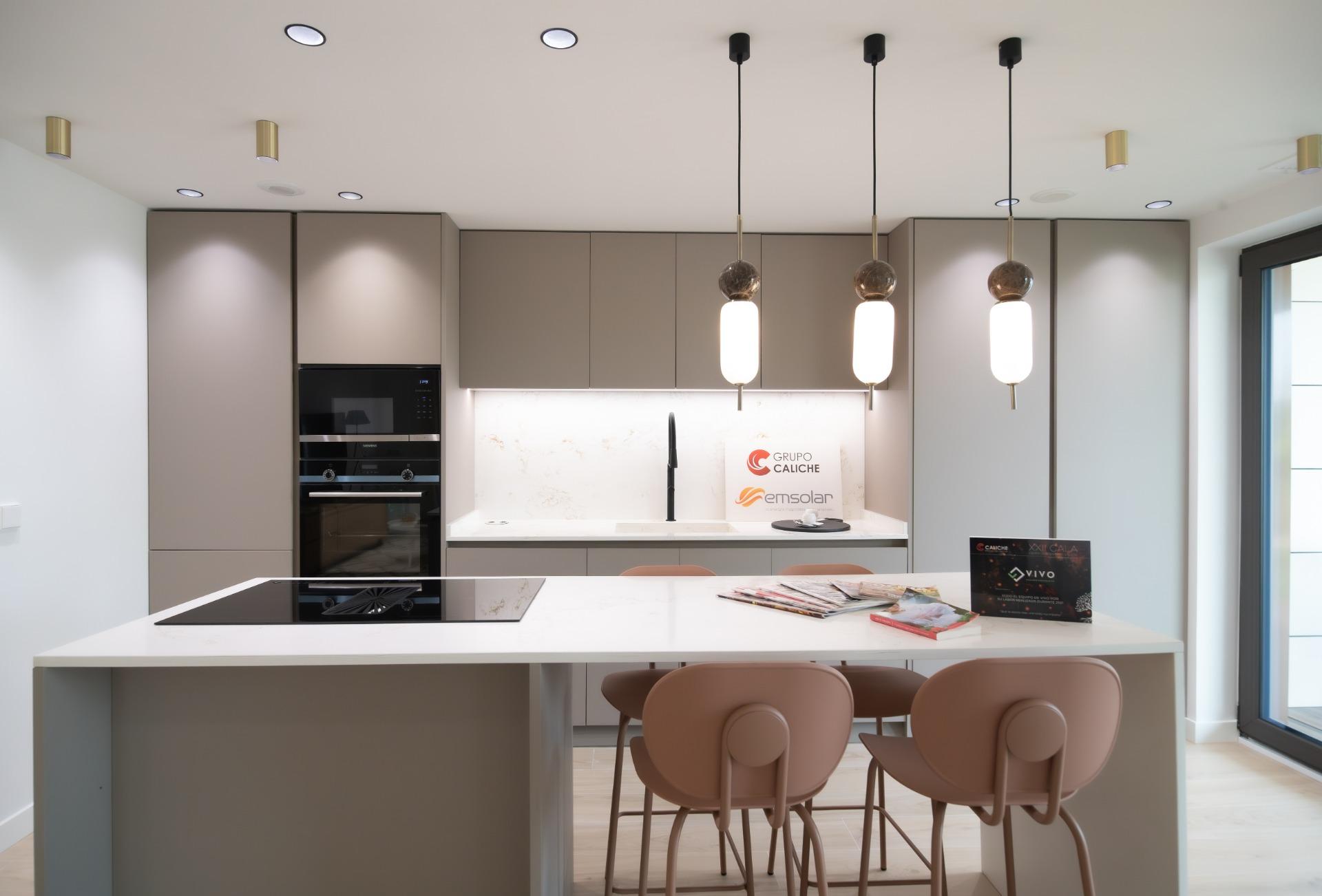 Modern kitchen with gray cabinets, built-in oven, marble backsplash, and a white island with four pinkish-brown stools beneath hanging pendant lights.