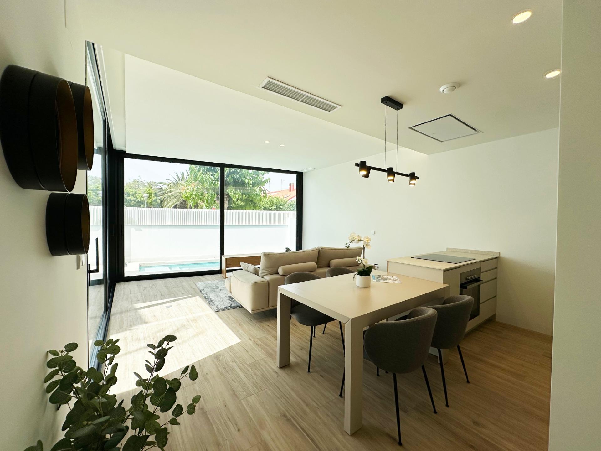 Modern open-plan living and dining area with a beige sofa, white dining table and gray chairs, large sliding glass doors to a courtyard or pool area, and a wall with circular decorative shelves on the left.