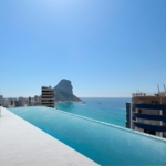Rooftop pool terrace with white tiles and lounge chairs, overlooking the blue sea and distant rocky island.