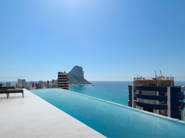 Rooftop pool terrace with white tiles and lounge chairs, overlooking the blue sea and distant rocky island.