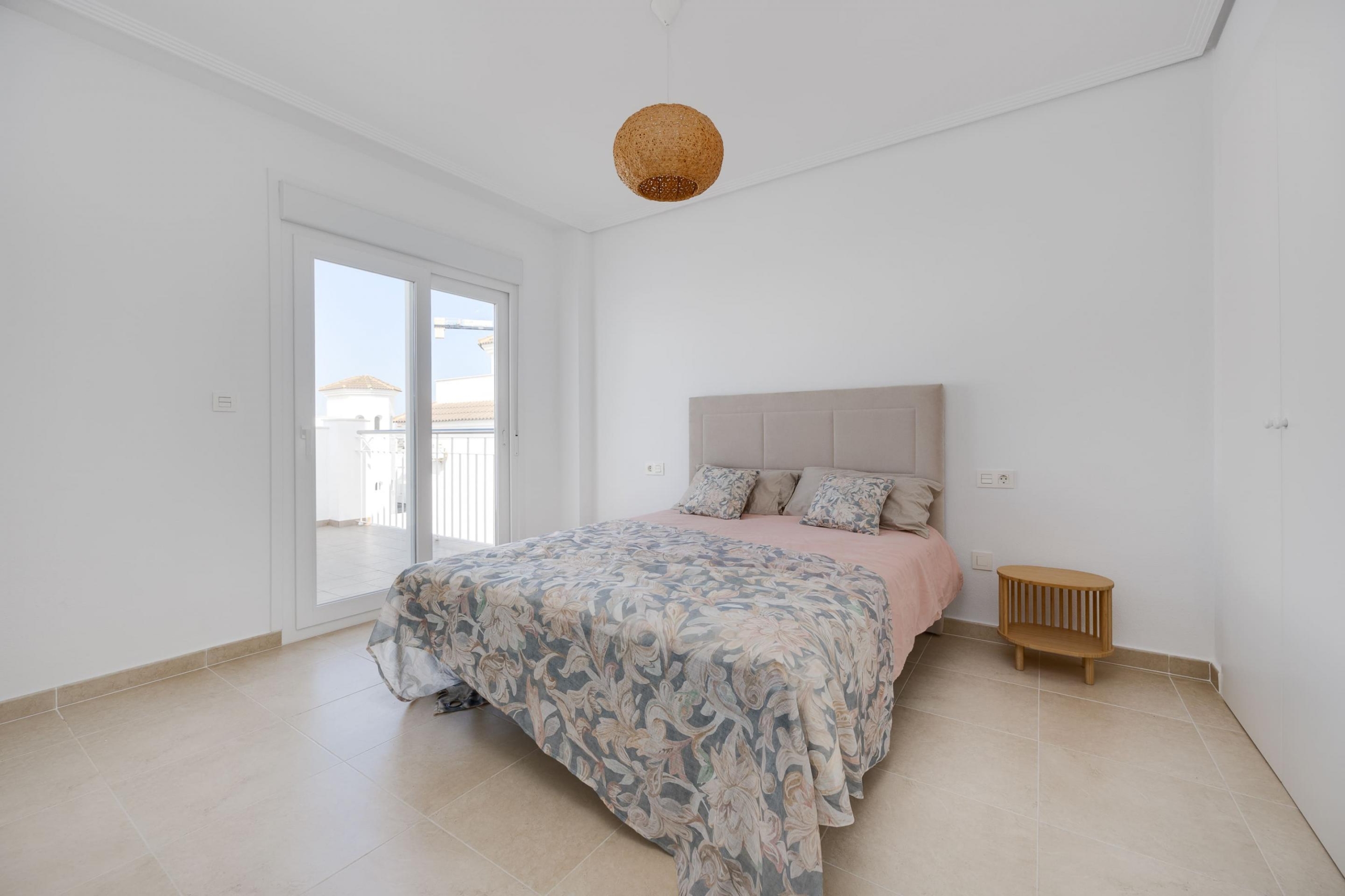 Bright bedroom with a double bed, beige headboard, floral quilt, and a woven pendant light hanging from the ceiling, near a balcony door to the left.