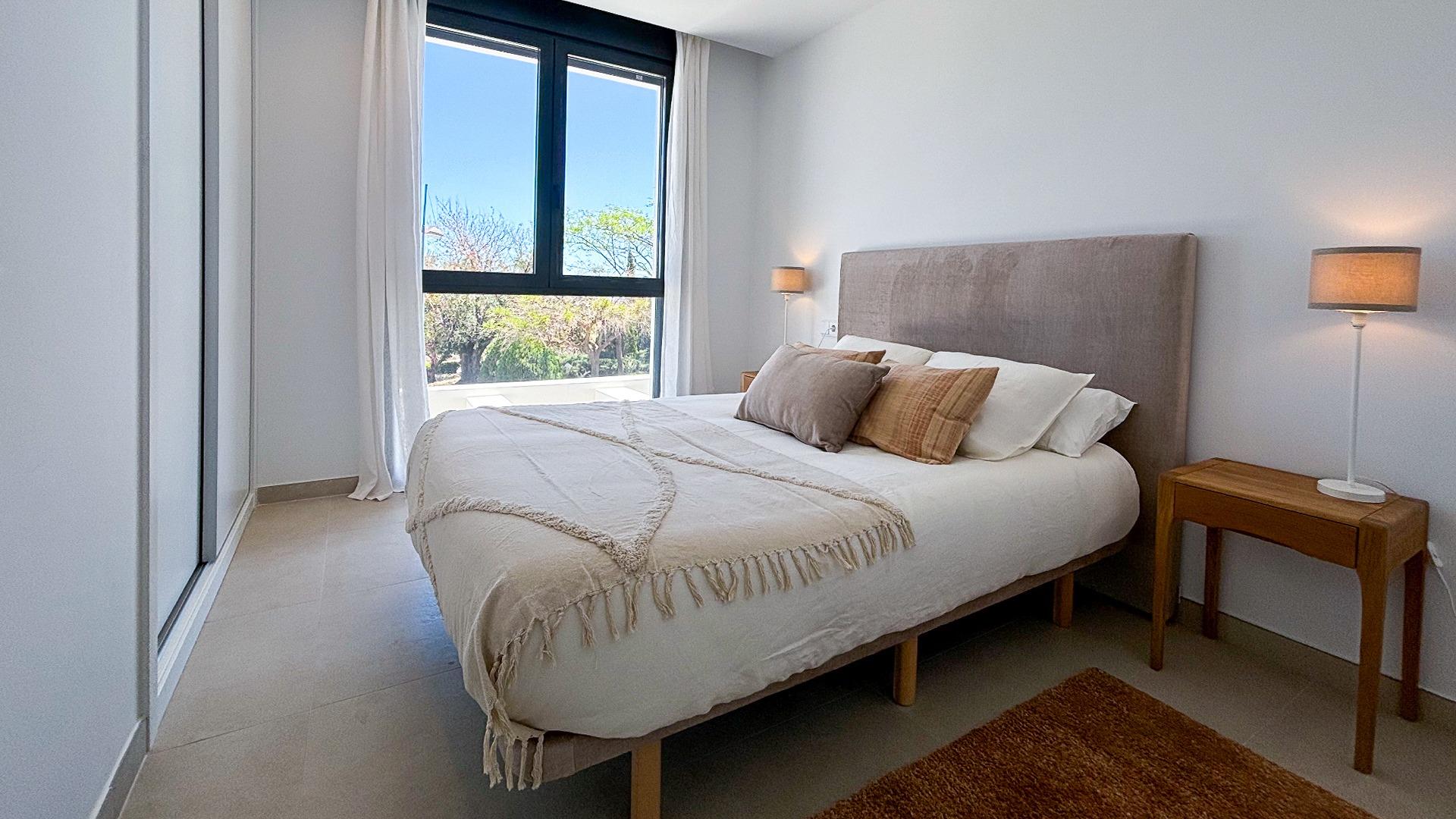 Bright bedroom with a padded beige headboard, white bedding, and a wooden nightstand with a lamp on the right. A large window brings in natural light with white curtains.
