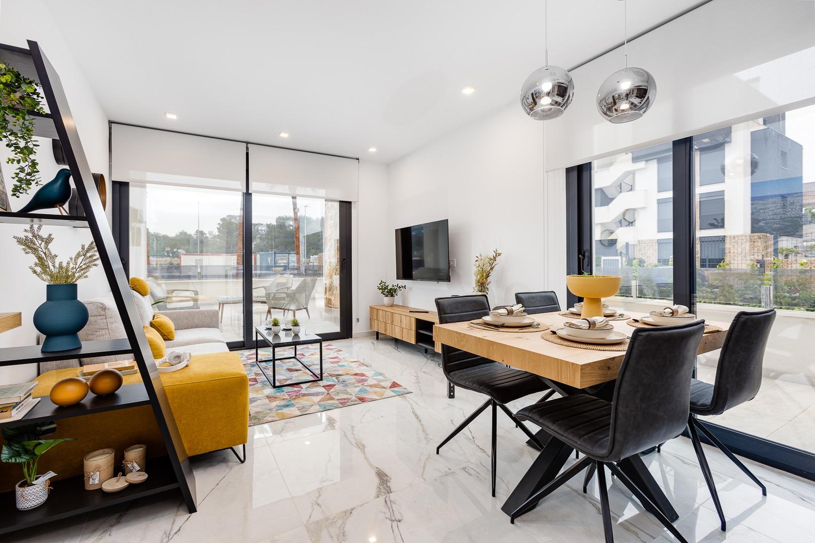 Open-plan living and dining room with a wooden dining table, black chairs, and a wall-mounted TV, bright with large windows.