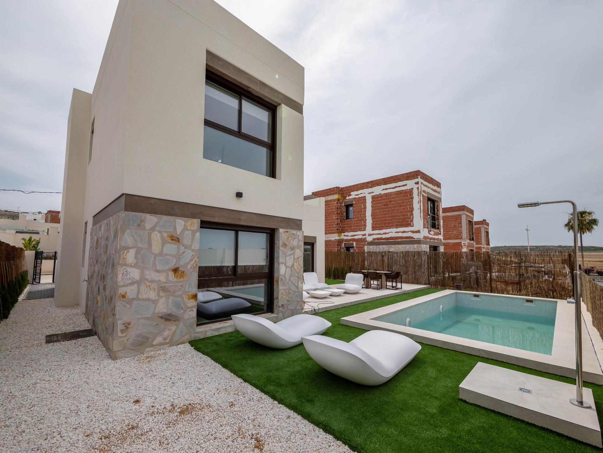 Modern villa with a rectangular in-ground pool, white curved lounge chairs, and an outdoor seating area along a stone and beige facade.