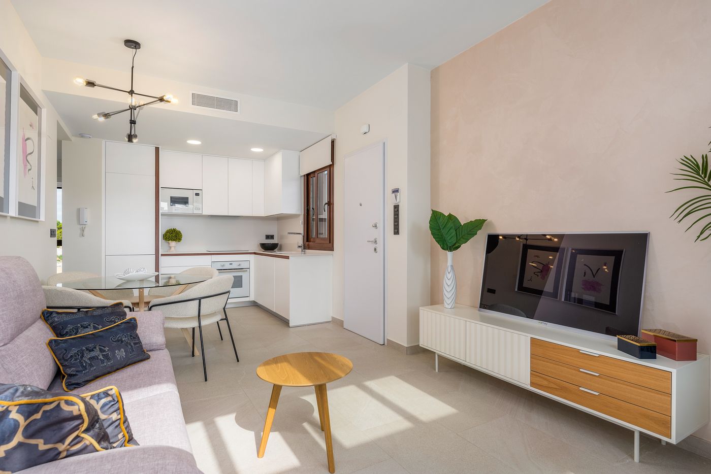 Open-concept living space with a white kitchen, light furniture, and a wall-mounted TV on a white-and-wood console.
