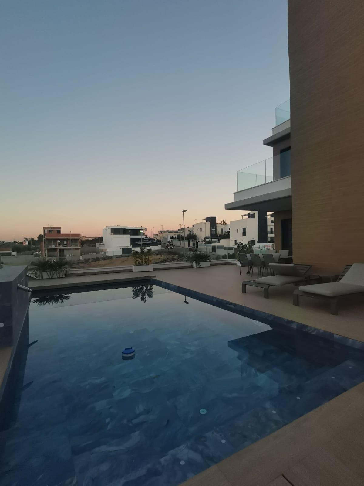 Modern apartment complex with a blue swimming pool and lounge chairs overlooking a calm sunset sky.