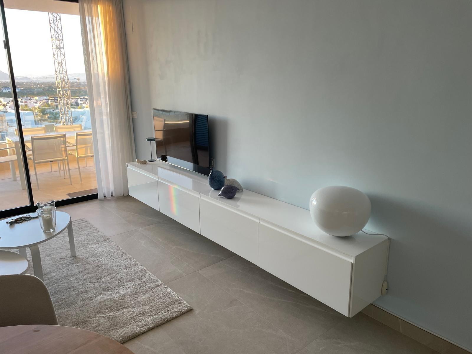 Modern living room with a low white media console, TV, and a large round white lamp along a pale blue wall; glass balcony doors on the left.
