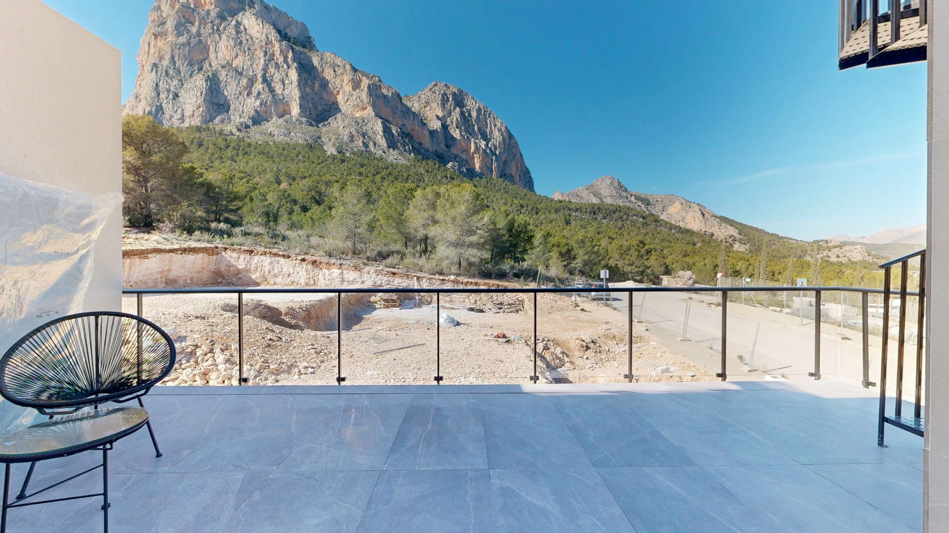 Balcony with glass railing overlooking a rocky mountain range and forest under a clear blue sky.