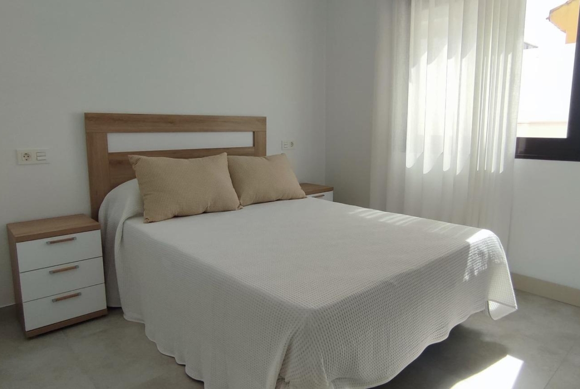 Double bed with a white textured bedspread and two beige throw pillows, wooden headboard and matching nightstand in a bright, minimalist bedroom with sheer curtains by a window on the right