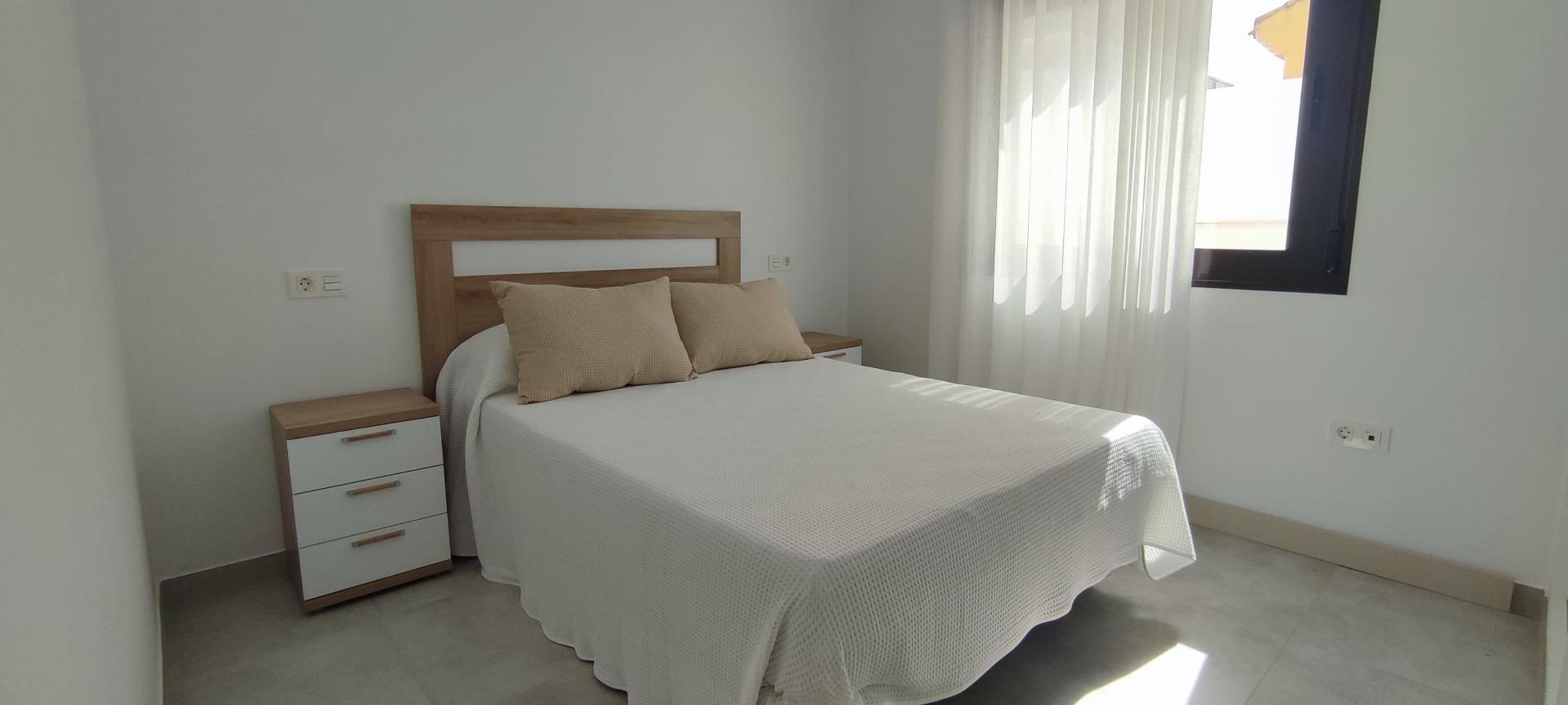 Double bed with a white textured bedspread and two beige throw pillows, wooden headboard and matching nightstand in a bright, minimalist bedroom with sheer curtains by a window on the right