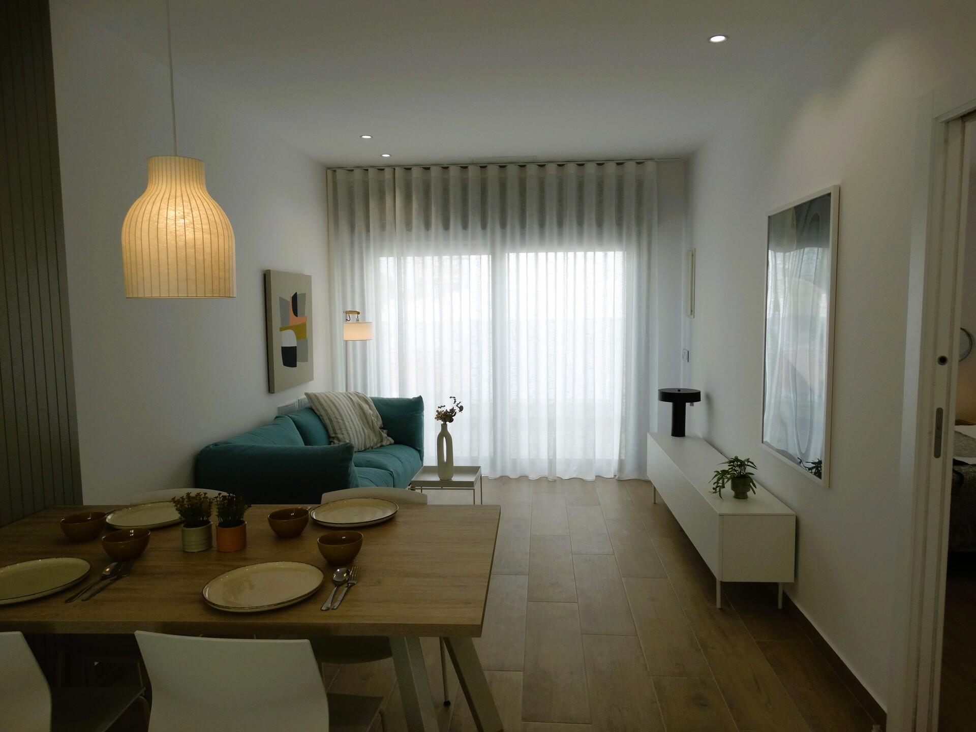Open-plan living/dining area with a wooden table set for six, a teal sofa, abstract wall art, and a large window with sheer white curtains -- bright and modern.