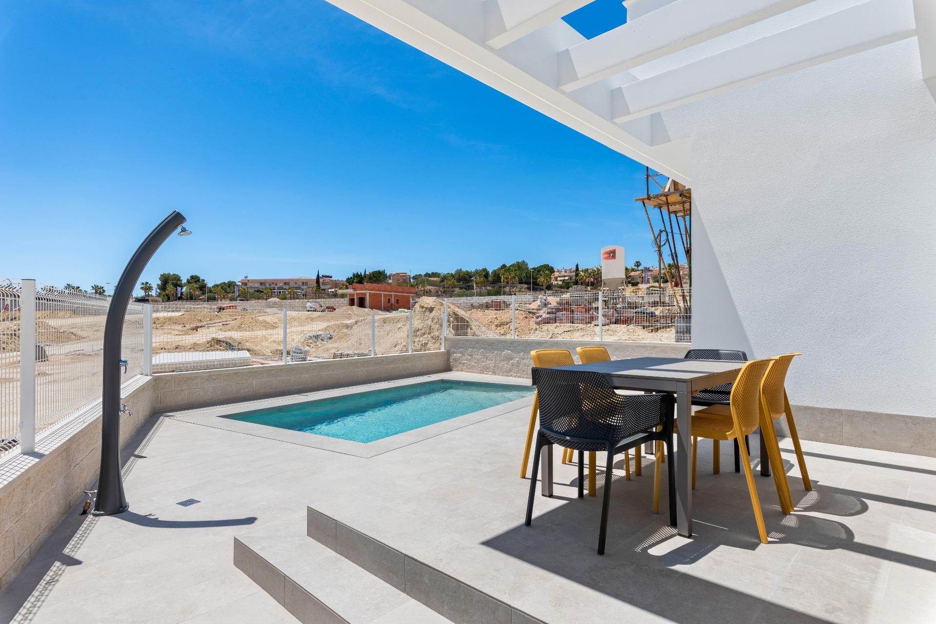 Patio with a small rectangular pool, a gray dining table and yellow chairs, white beams overhead, and a construction site beyond under a blue sky.