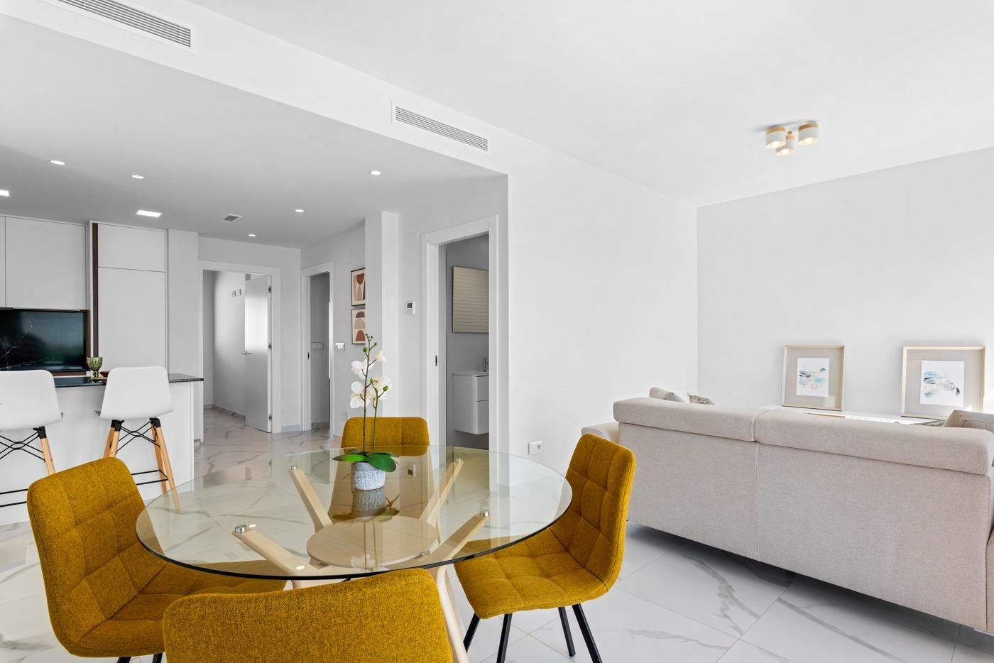 Open-plan living and dining area with a round glass table, mustard chairs, beige sofa, and marble tile floor.