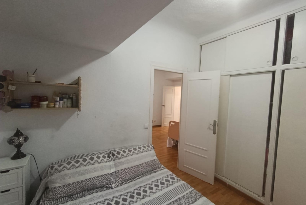 Bedroom with white walls and built-in wardrobes, a bed with gray geometric bedding, a shelf of toiletries, and an open door to a hallway with wooden floor.