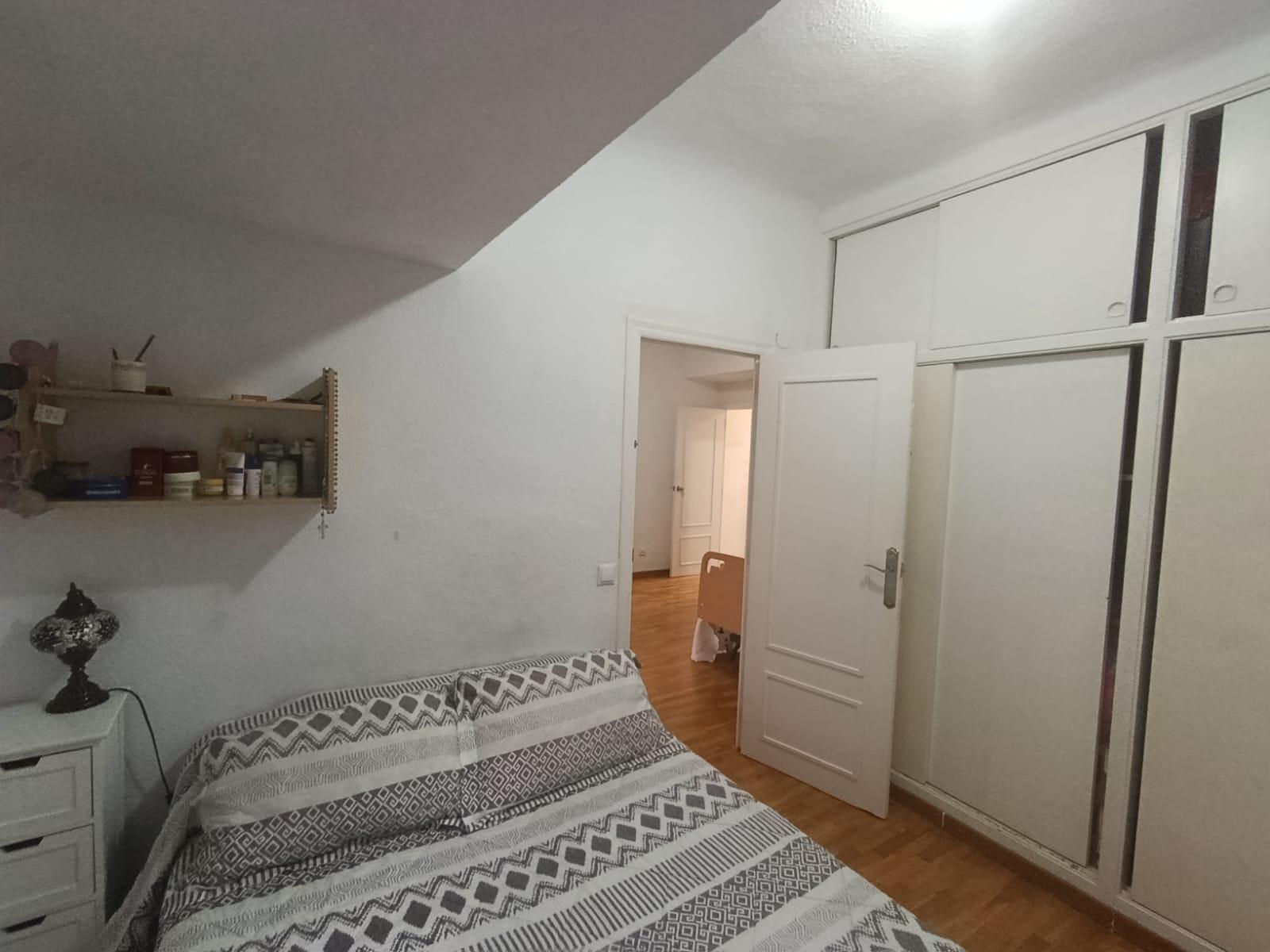 Bedroom with white walls and built-in wardrobes, a bed with gray geometric bedding, a shelf of toiletries, and an open door to a hallway with wooden floor.
