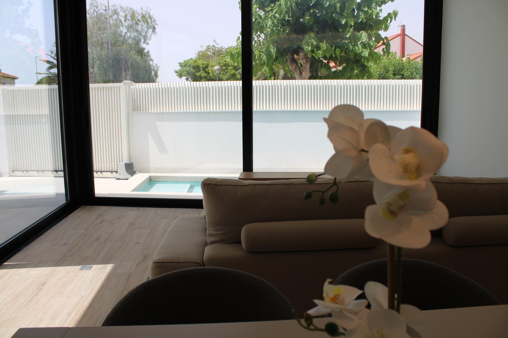 Living room with a beige sofa, large glass doors, and white orchid flowers in the foreground; view of a fenced yard with a small pool and trees outside.”