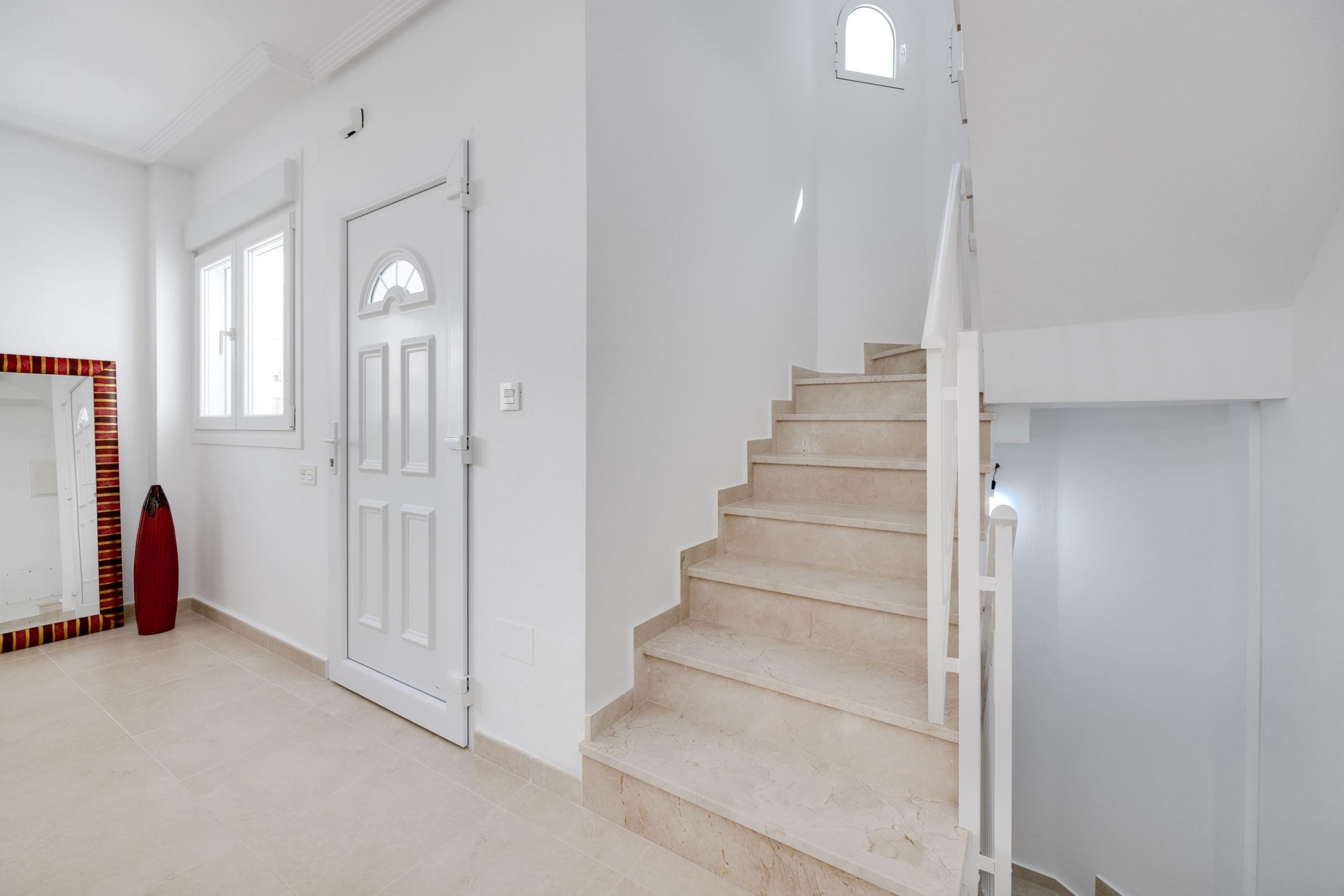 White entryway with beige marble stairs to the right, a white door with an arched window, and a tall red vase beside a striped mirror on the left wall.