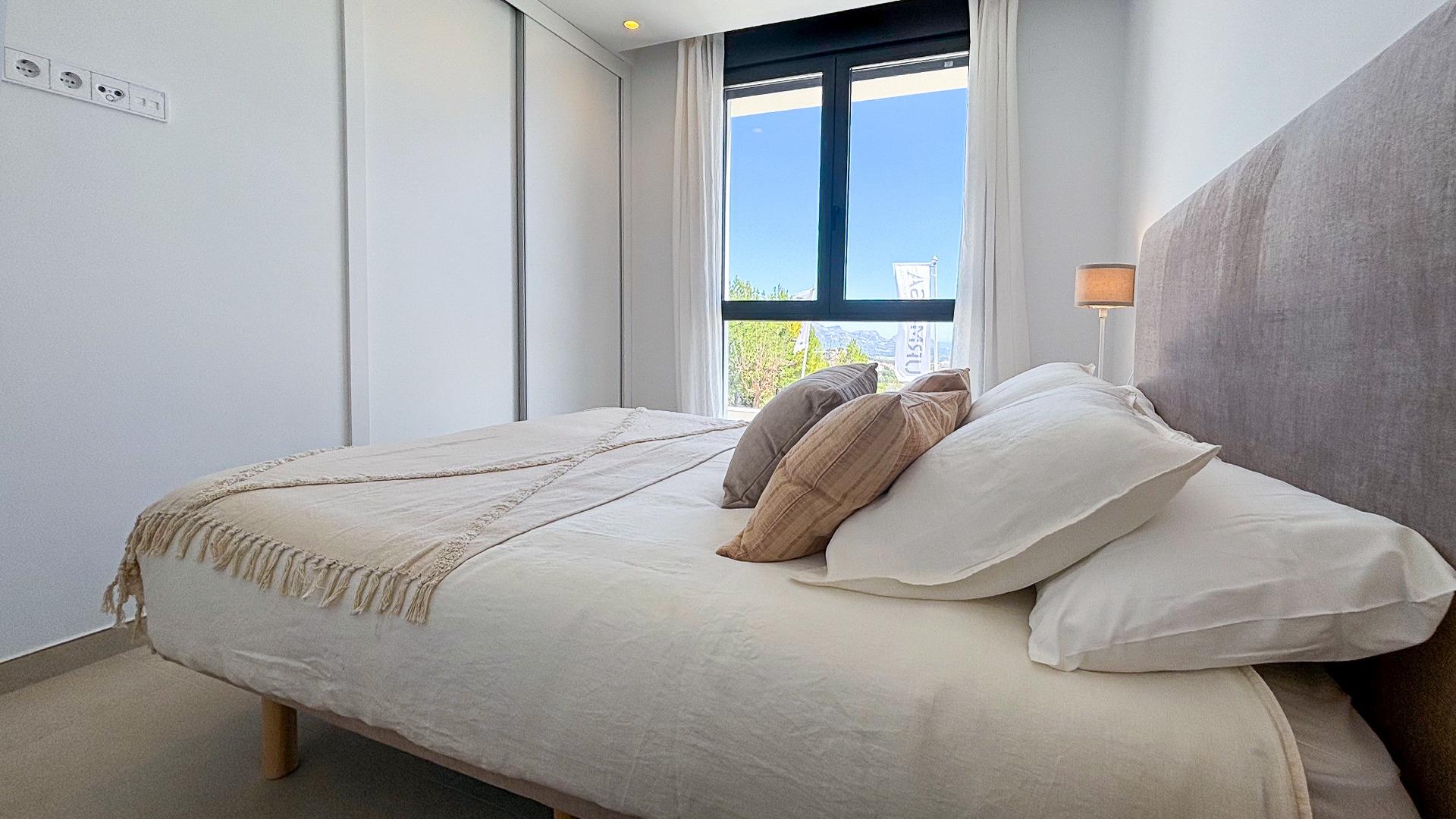 Bright, modern bedroom with a white bed, beige throw and multiple pillows, a tall headboard, and a large window with white curtains showing a blue sky outside.