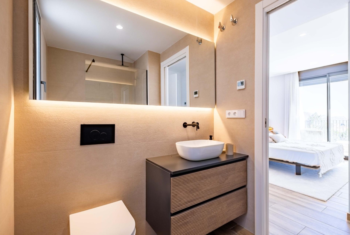 Modern bathroom with a white vessel sink on a dark wood vanity and a large backlit mirror; doorway opens to a bright bedroom.